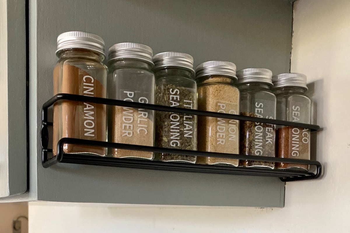 A spice rack holds clear glass jars labeled with various seasonings, including cinnamon, garlic powder, Italian seasoning, onion powder, and sea seasoning. The jars are displayed on a metal rack mounted on a light-colored wall, providing easy access for cooking.