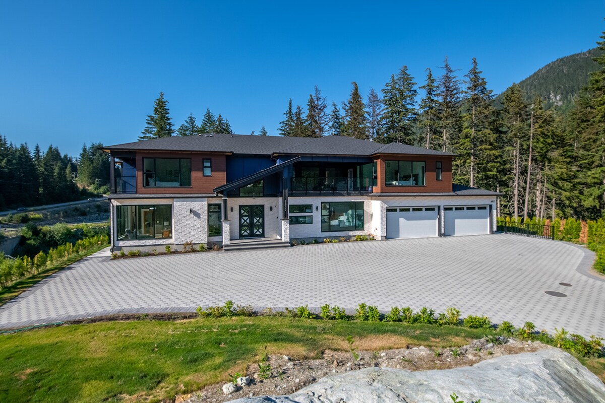 A modern two-story home is set against a backdrop of trees, with a large stone driveway in front. Expansive windows reflect the blue sky, and the building features a mix of dark and light exterior materials. The three-car garage is included, providing ample parking space.