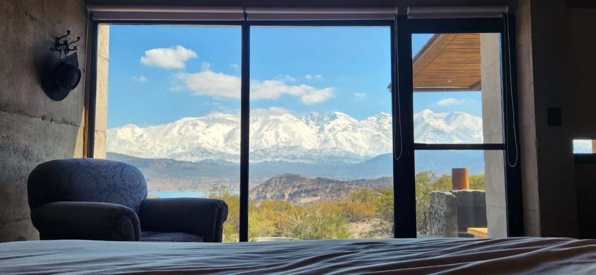 A spacious bedroom showcases a king-sized bed with a view through large windows, revealing the snow-capped Andes Mountains in the distance. A comfortable armchair sits beside the window, complementing the serene landscape.