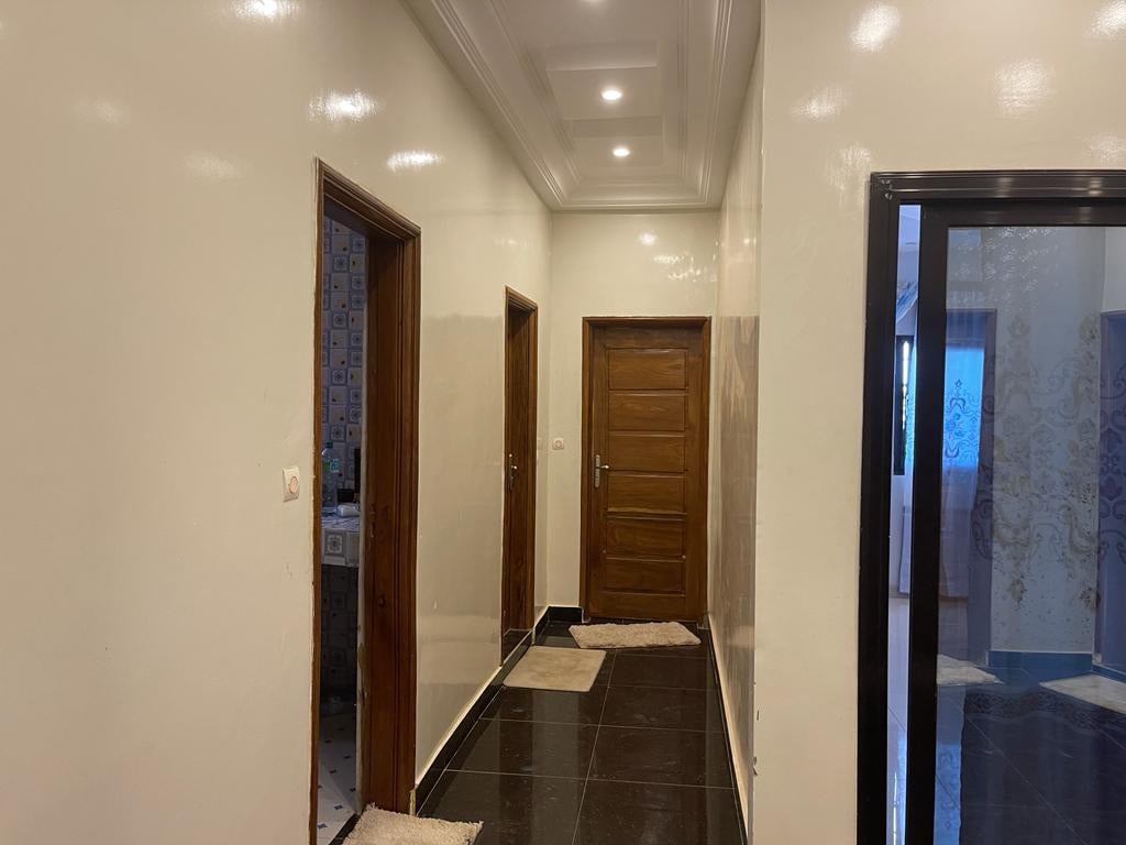 A hallway is seen, featuring glossy white walls and dark tiled flooring. Each door offers access to different rooms, with one door leading to a bathroom visible on the left. Soft lighting illuminates the space, enhancing the clean and open feel.