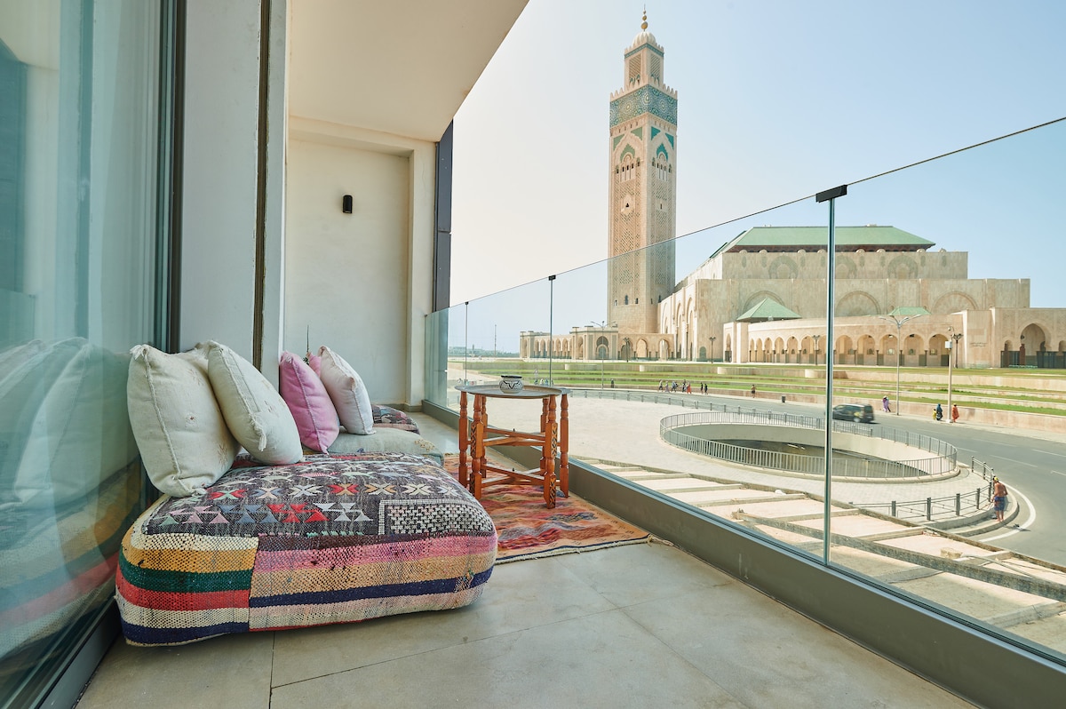 An outdoor terrace featuring a colorful cushioned seating area overlooks the Mosquée Hassan II. Decorative pillows are arranged on a large, textured mat, while a small wooden table complements the setting. Clear glass railings provide an unobstructed view of the surrounding area.
