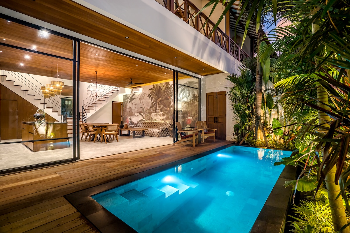 A private pool is visible in the foreground, surrounded by lush greenery and wooden decking. The villa's spacious interior is partially seen through expansive glass doors, highlighting a warmly lit living area with comfortable seating and dining space, accented by tropical artwork and elegant lighting.