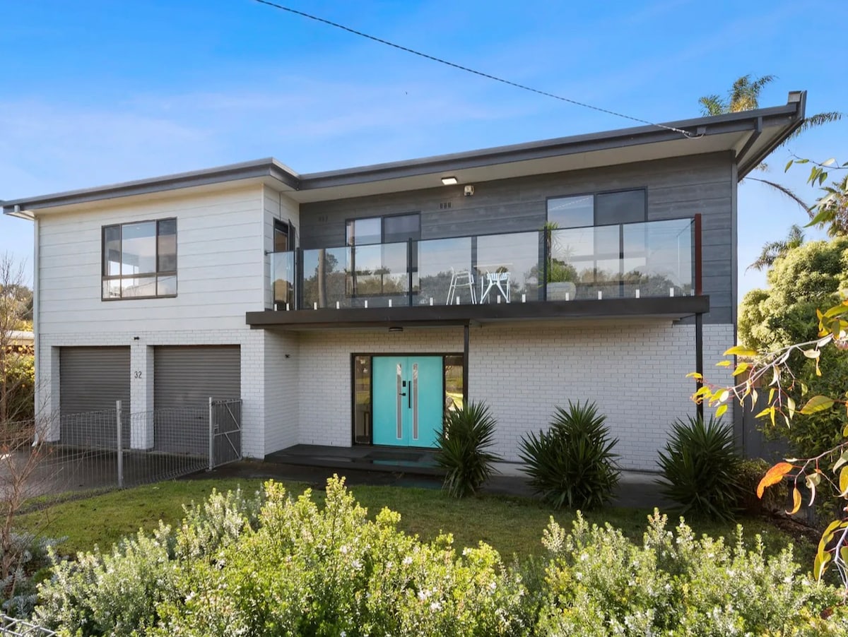 The exterior of a two-story home is shown, featuring a contemporary design with large windows and a spacious balcony. A vibrant turquoise front door welcomes guests, while lush greenery surrounds the property, adding to its inviting presence.