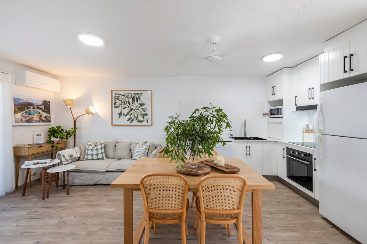 An open-plan living area showcases a light-colored couch and a wooden dining table with rattan chairs. Modern kitchen appliances are visible, alongside cabinetry in a clean white finish. Natural light filters through the window, complemented by decorative plants and artwork.