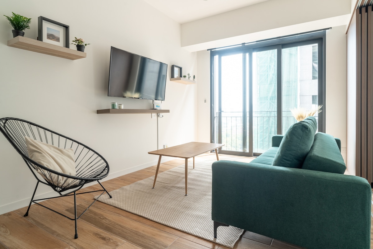 A modern living area features a comfortable green sofa and a stylish lounge chair. A wooden coffee table is positioned on a textured rug. Natural light fills the space through large glass doors leading to a balcony, enhancing the open atmosphere.