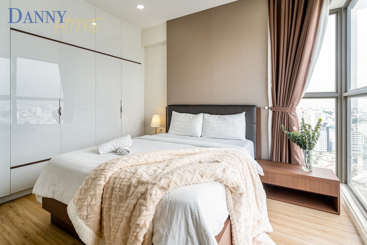 A minimalist bedroom features a spacious bed dressed in crisp white linens, complemented by a soft beige throw. A bedside table holds a small lamp and a vase with fresh greenery. Large windows provide natural light, framed by soft, brown curtains.