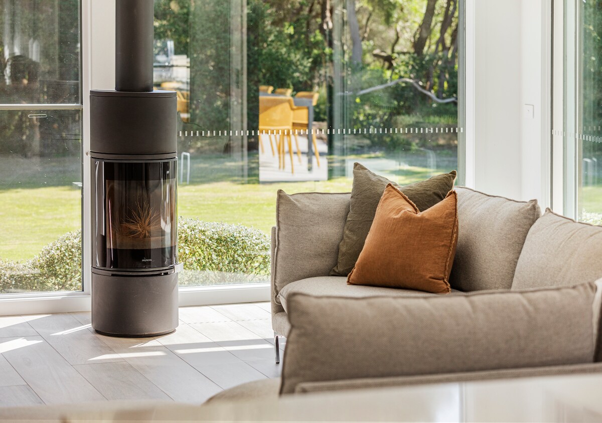 A modern living space features a sleek, circular fireplace as a focal point, surrounded by large glass windows that provide a view of the lush outdoor garden. A cozy sofa with plush cushions adds to the inviting atmosphere.