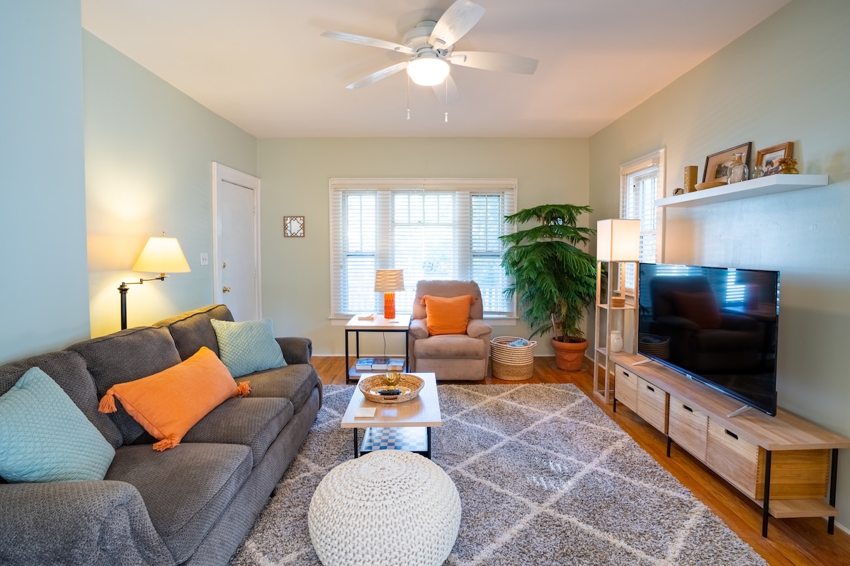 A cozy living room is presented with a sectional sofa and a single armchair, adorned with colorful throw pillows. Natural light filters through multiple windows, highlighting the neutral wall colors. A large flat-screen TV is positioned on a wooden console, complemented by a decorative coffee table.