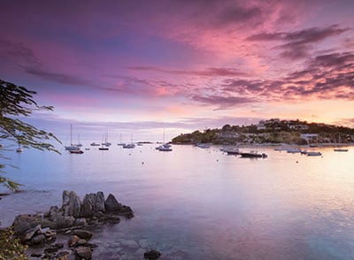 The serene cove features calm waters reflecting hues of pink and purple from the sunset. Several boats are anchored, while rocky outcrops are visible along the shoreline, framed by soft greenery. A peaceful atmosphere is established by the surrounding scenery.