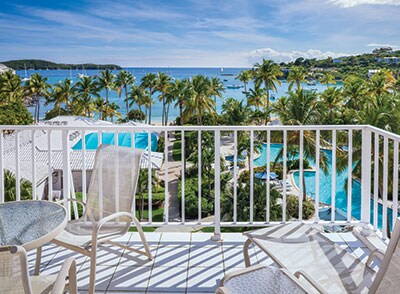 A balcony view displays a shimmering blue ocean framed by palm trees. Several boats are anchored in the water, while two swimming pools are visible within the resort grounds, offering a peaceful atmosphere for relaxation.