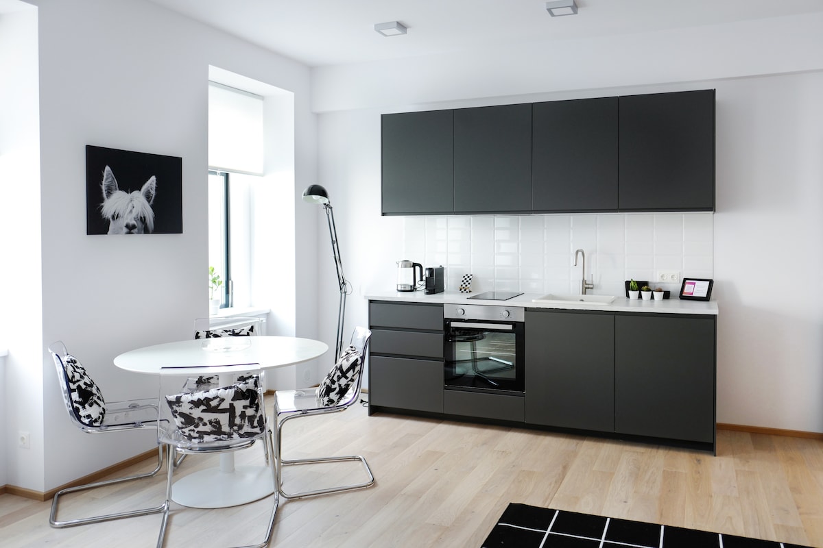 A modern kitchen area features sleek cabinetry in dark tones and a minimalist design. A round dining table with distinctive patterned chairs is positioned near a large window, allowing natural light to brighten the space. Essential appliances, including a coffee machine, are visibly arranged.