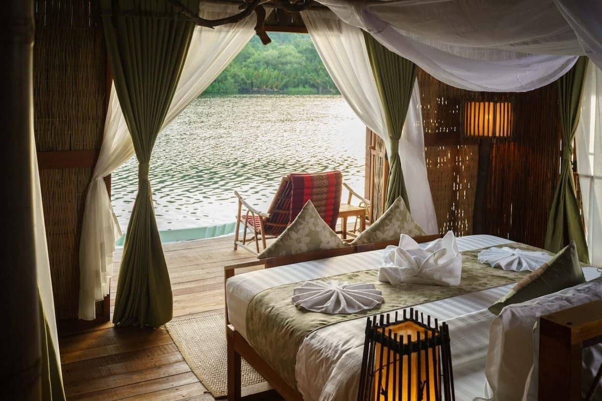 A serene bedroom features a spacious bed adorned with decorative towels and a light-colored blanket. Natural light floods the room through flowing curtains, leading to a private balcony with a comfortable chair and a blanket draped over it. A calm water view is visible beyond.