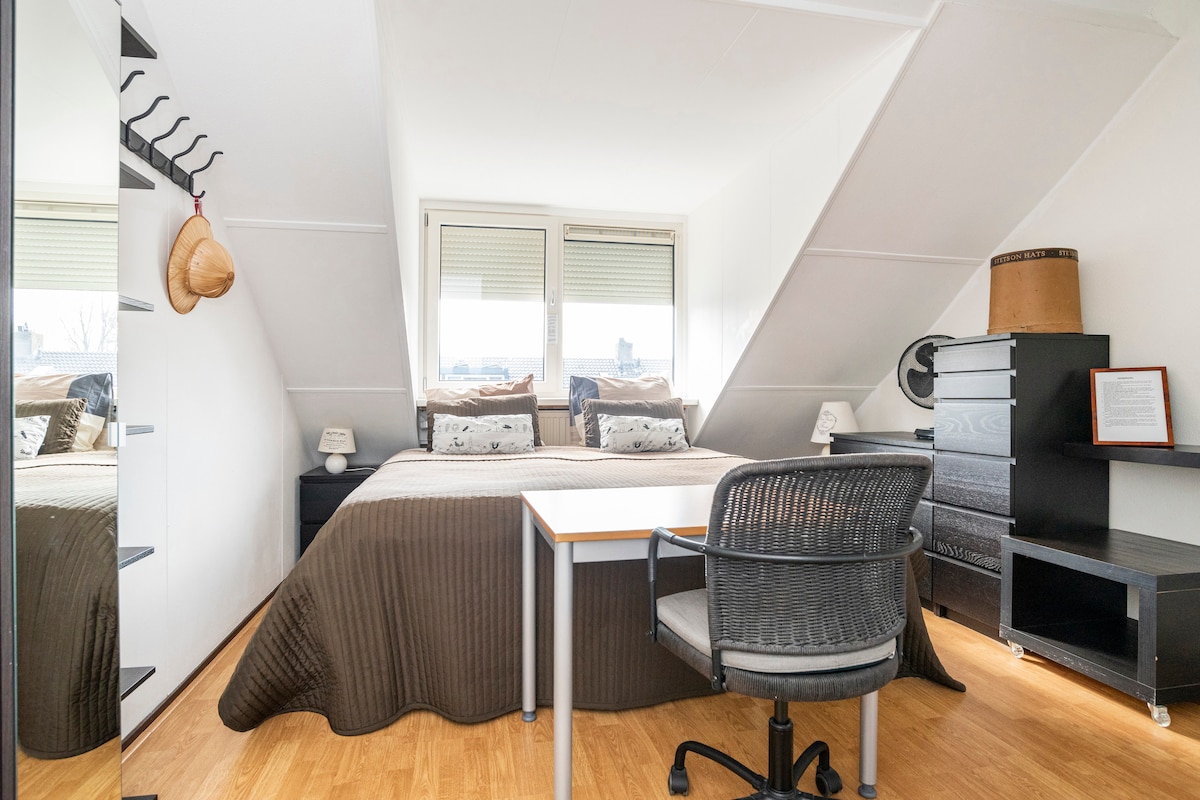 A cozy bedroom is visible, featuring a spacious bed with a neutral quilt. Natural light enters through large windows, illuminating a small desk with an office chair. A wardrobe and a few decorative items complement the warm wooden flooring.