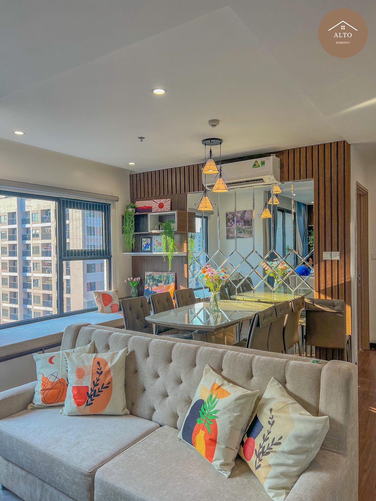 A well-furnished living area features a cozy sofa adorned with decorative pillows. A glass dining table is surrounded by colorful chairs and positioned near large windows, allowing natural light to illuminate the space. Decorative plants and artwork add a touch of character.