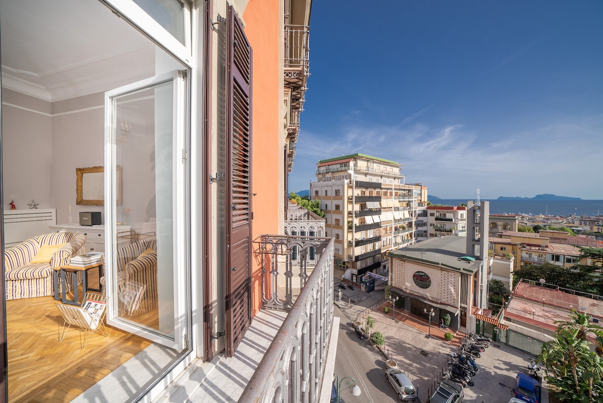 A balcony view reveals a vibrant neighborhood scene with buildings and palm trees. The urban landscape is framed by a clear blue sky and distant views of the coastline, showcasing the lively atmosphere of the surrounding area.