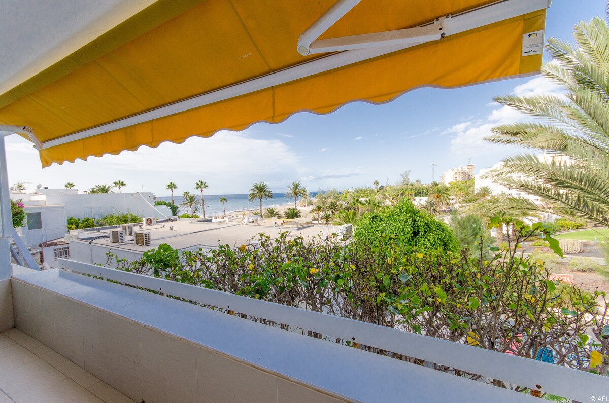 A balcony view features a bright yellow awning and lush greenery. The scene captures a glimpse of the sea, framed by palm trees. The surroundings include low-rise buildings and landscaped areas, creating a serene atmosphere.