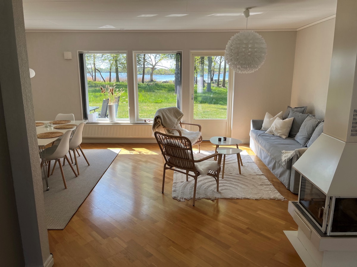 A light-filled living area features large windows offering views of the greenery and water outside. A cozy sofa and woven chairs surround a coffee table on warm wooden flooring. A dining table with modern chairs is set nearby, creating a welcoming space for relaxation.