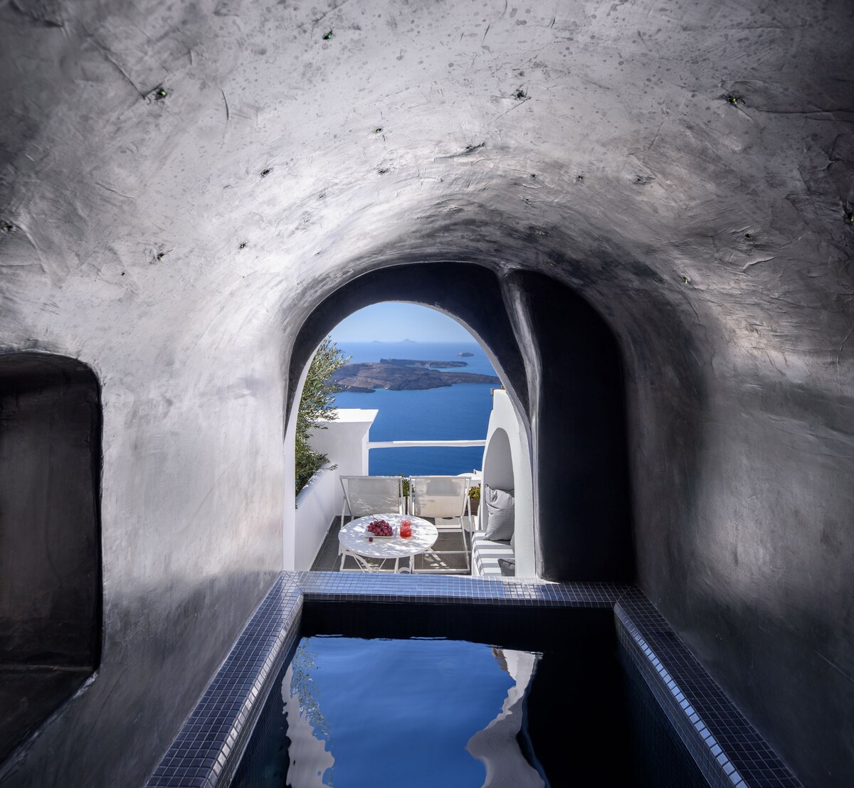 An arched entryway frames a private plunge pool with dark water, leading to a furnished terrace with stunning views of the caldera and Aegean Sea. Natural light illuminates the smooth curved walls, enhancing the serene atmosphere.