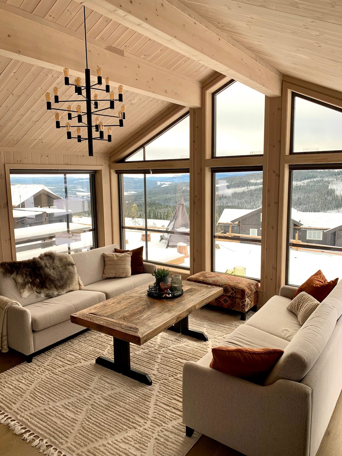 A bright living area features two light-colored sofas arranged around a rustic wooden table. Large windows provide views of snow-covered mountains and surrounding landscapes. A chandelier hangs overhead, and decorative pillows add a cozy touch to the space.