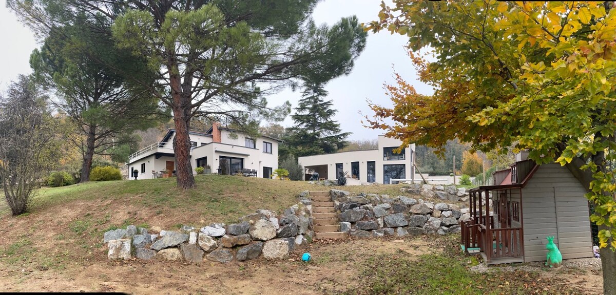 The exterior of the house is showcased, featuring a modern design with large windows. A grassy area is bordered by a stone wall and steps leading to the entrance. Trees and autumn foliage add natural elements, while a small playhouse is visible in the foreground.