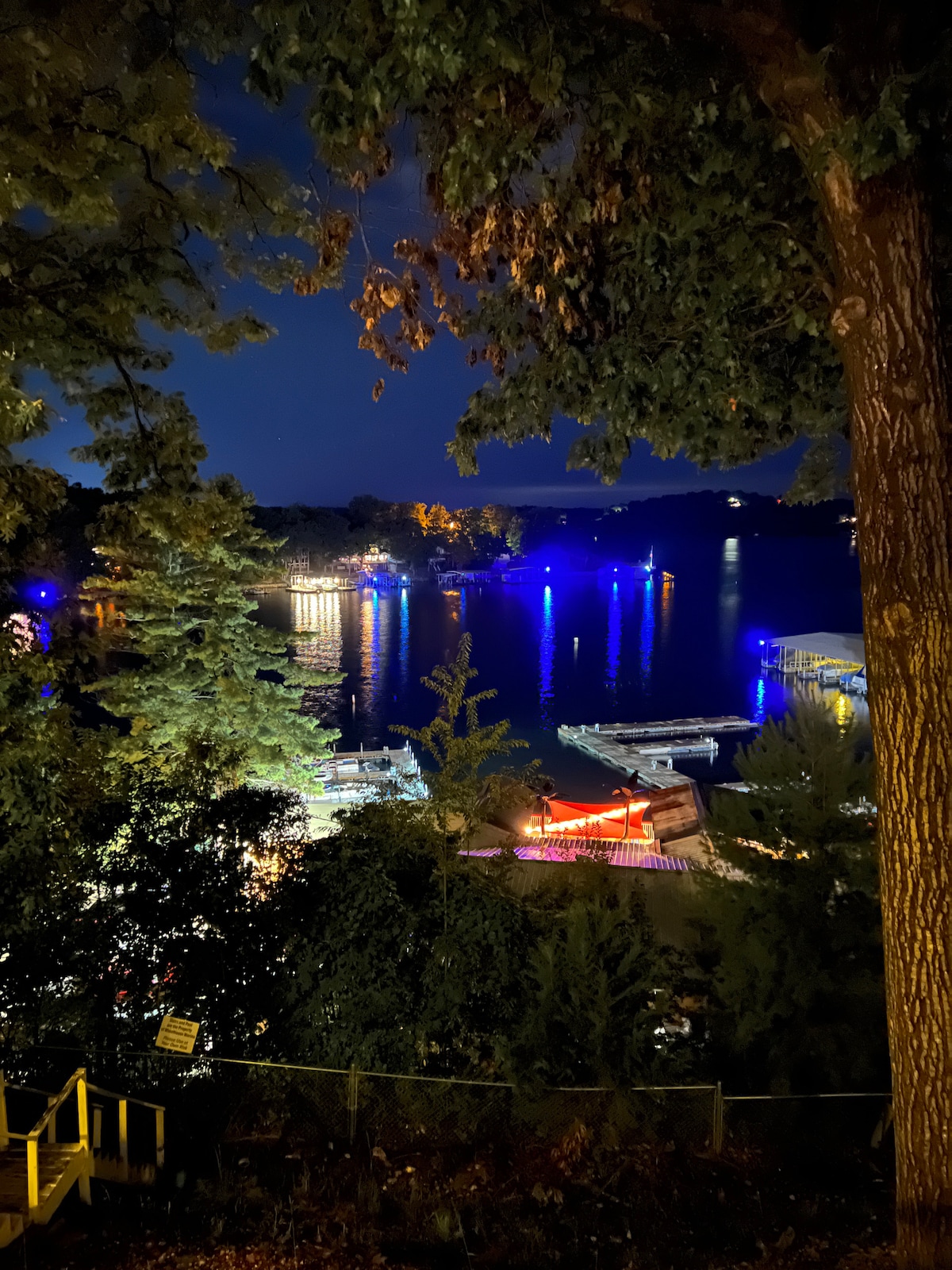 The scene captures a nighttime view of the lake, with boats docked along the shoreline. Soft blue and colorful lights reflect off the water, creating a serene atmosphere. Trees frame the image, adding depth and a touch of nature to the lakeside setting.