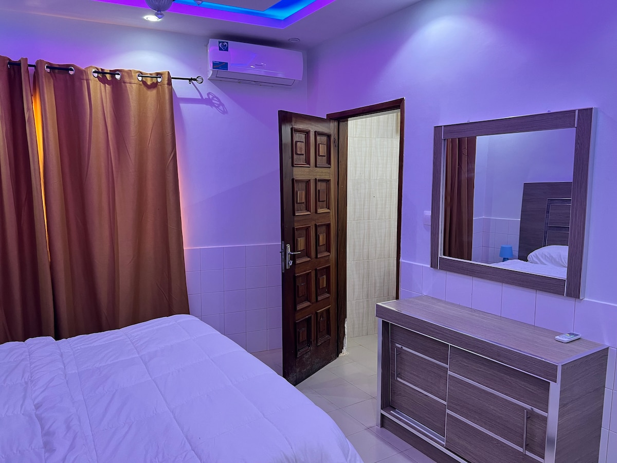A bright room features a spacious bed covered with a white quilt. Nearby, a large mirror is mounted above a wooden dresser. Soft brown curtains frame a window, and the doorway leads to a bathroom. Ambient lighting creates a calming atmosphere.