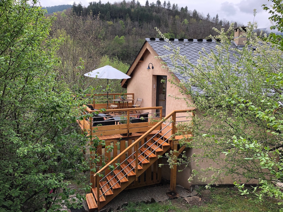 An exterior view captures the gîte surrounded by lush greenery, featuring a wooden deck accessed by a staircase. A patio umbrella provides shade over the outdoor seating area, ideal for relaxation in nature. The structure displays a rustic facade with a slate roof.