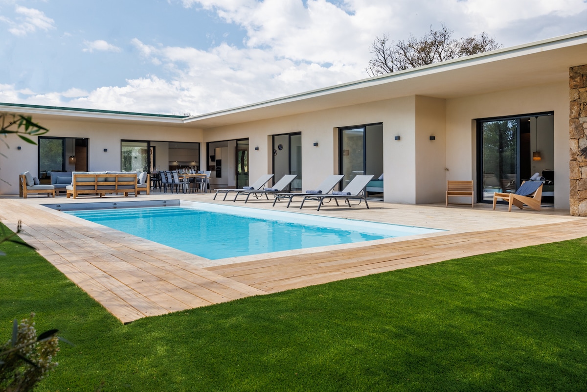 The exterior of the villa features a heated pool surrounded by a wooden deck. Comfortable loungers are positioned nearby. A spacious terrace extends from the main living area, providing access to the lush green lawn and large glass doors leading into the interior.