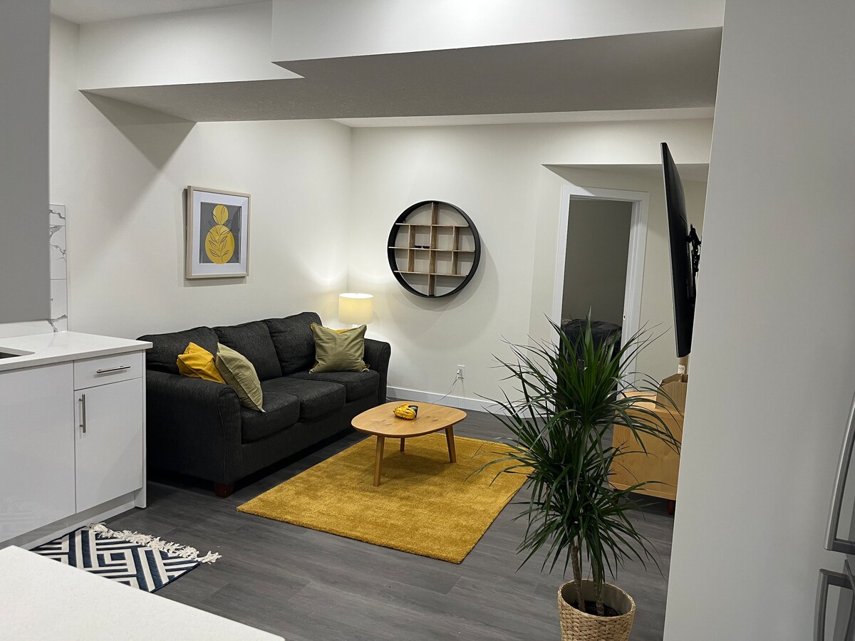 A welcoming living area is featured, with a dark sofa adorned with green cushions and a round coffee table. A vibrant yellow rug is positioned beneath the coffee table. A wall-mounted TV provides entertainment, while a plant in a decorative pot adds a touch of greenery.