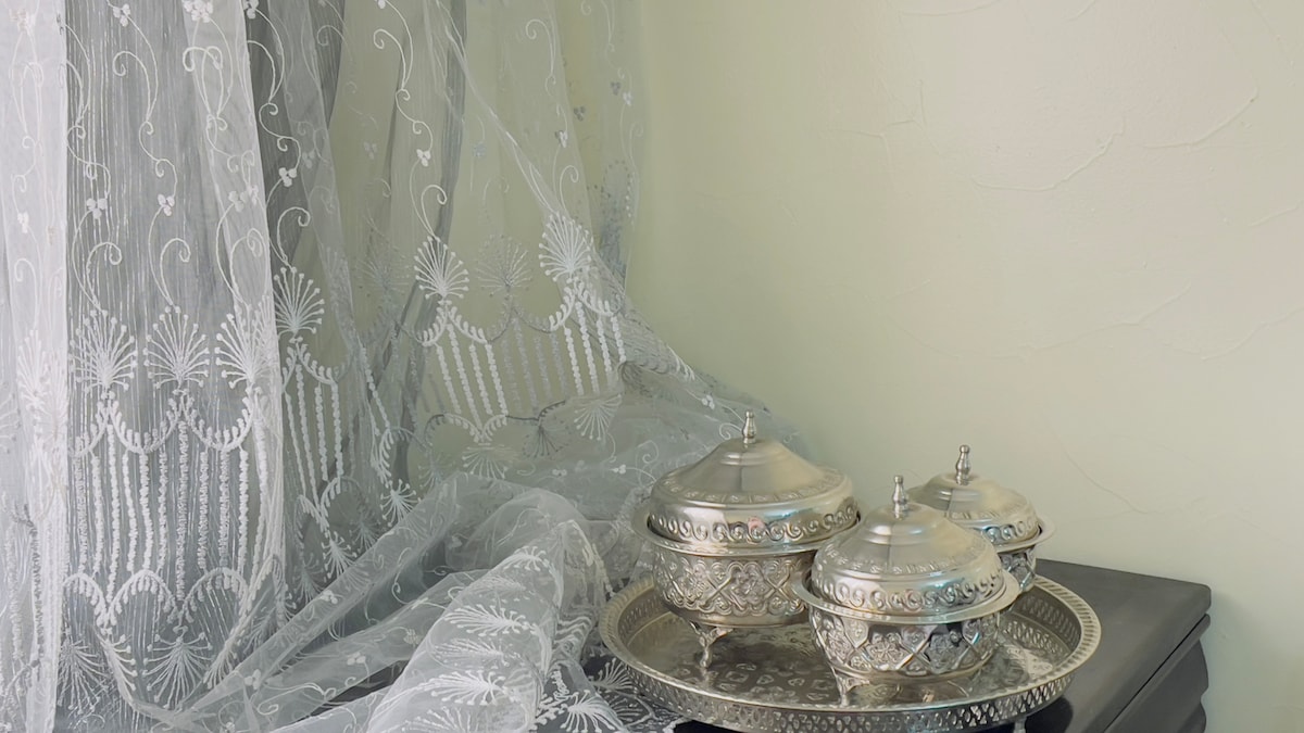 Intricately designed sheer curtains drape gracefully beside a decorative silver tray. Three ornate silver bowls are arranged on the tray, reflecting light softly. The walls are painted in a neutral tone, providing a calming backdrop for the elegant decor.