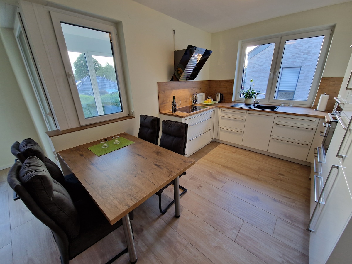 A modern kitchen is seen featuring sleek cabinetry and a wooden dining table with four upholstered chairs. Natural light enters through large windows, illuminating the countertops and the integrated cooking area. A potted plant adds a touch of greenery to the space.