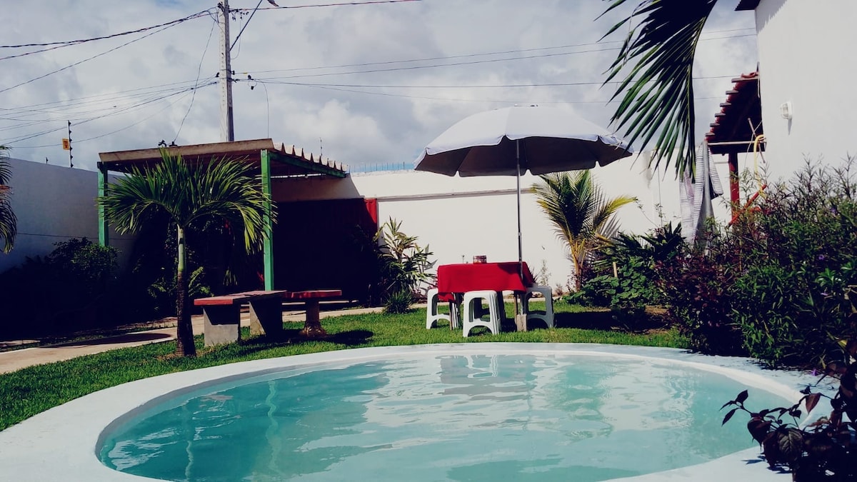A circular swimming pool is visible, surrounded by a well-maintained grassy area. A red tablecloth covers a table with white chairs under a large umbrella. Tropical plants and a wooden bench are also present, contributing to the outdoor relaxation space.
