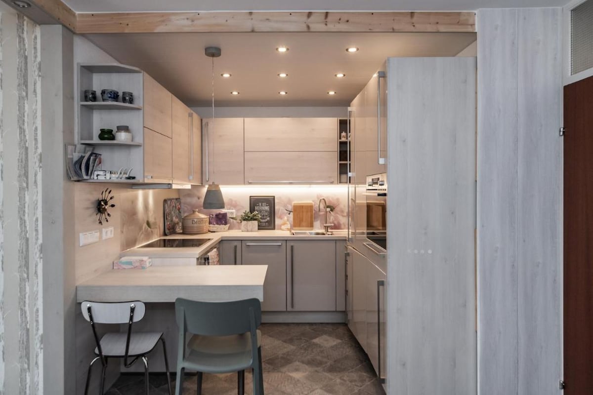 A modern kitchen is presented with sleek cabinetry and a light wood finish. Subtle lighting is provided by recessed ceiling fixtures. A small dining table is paired with minimalist chairs, while artwork and kitchen accessories enhance the functional space.