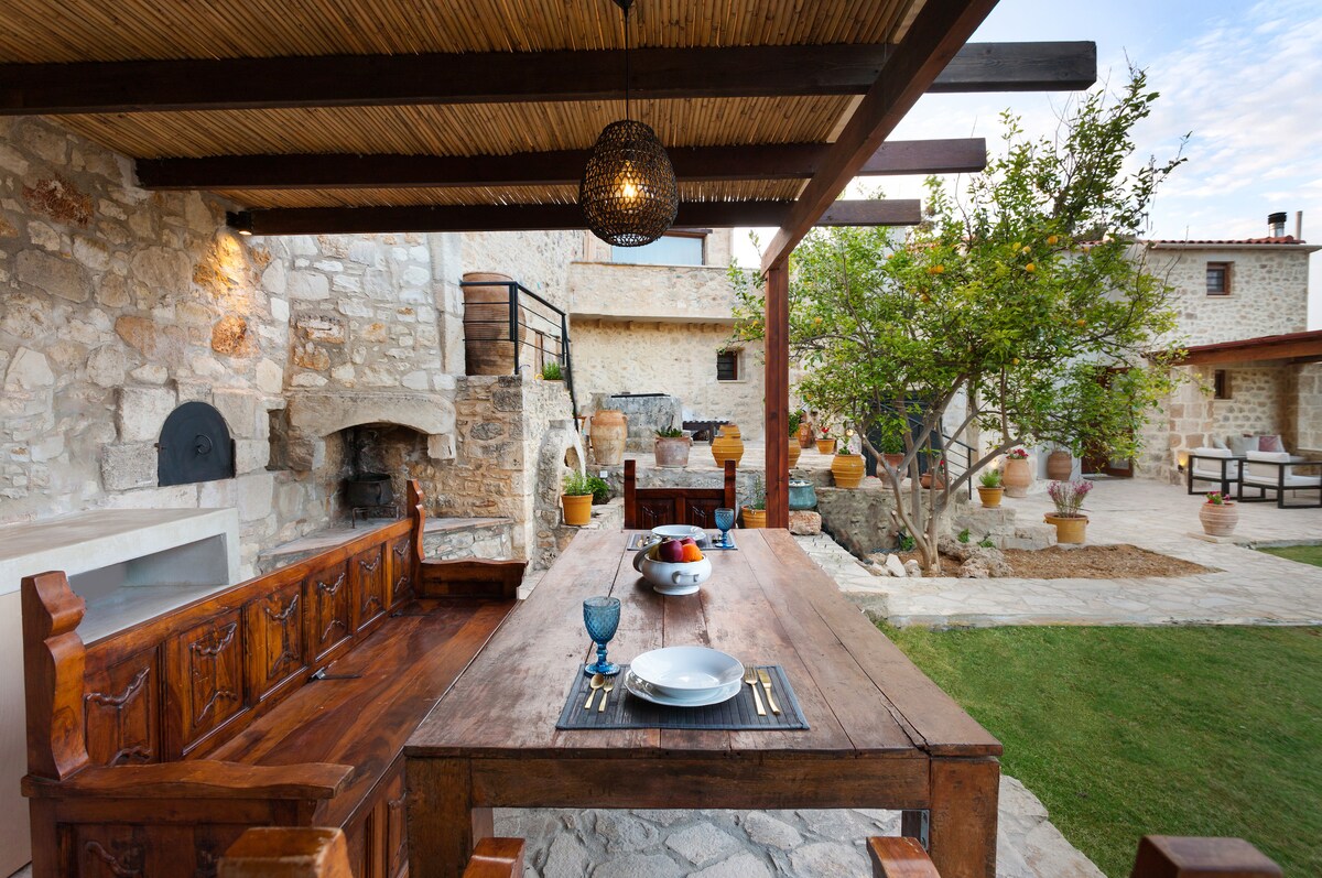 An outdoor dining area is presented under a shaded pergola, featuring a rustic wooden table set for a meal. Stone walls surround the space, and a garden with greenery and pots adds a natural touch. An outdoor barbecue and seating areas are visible in the background.