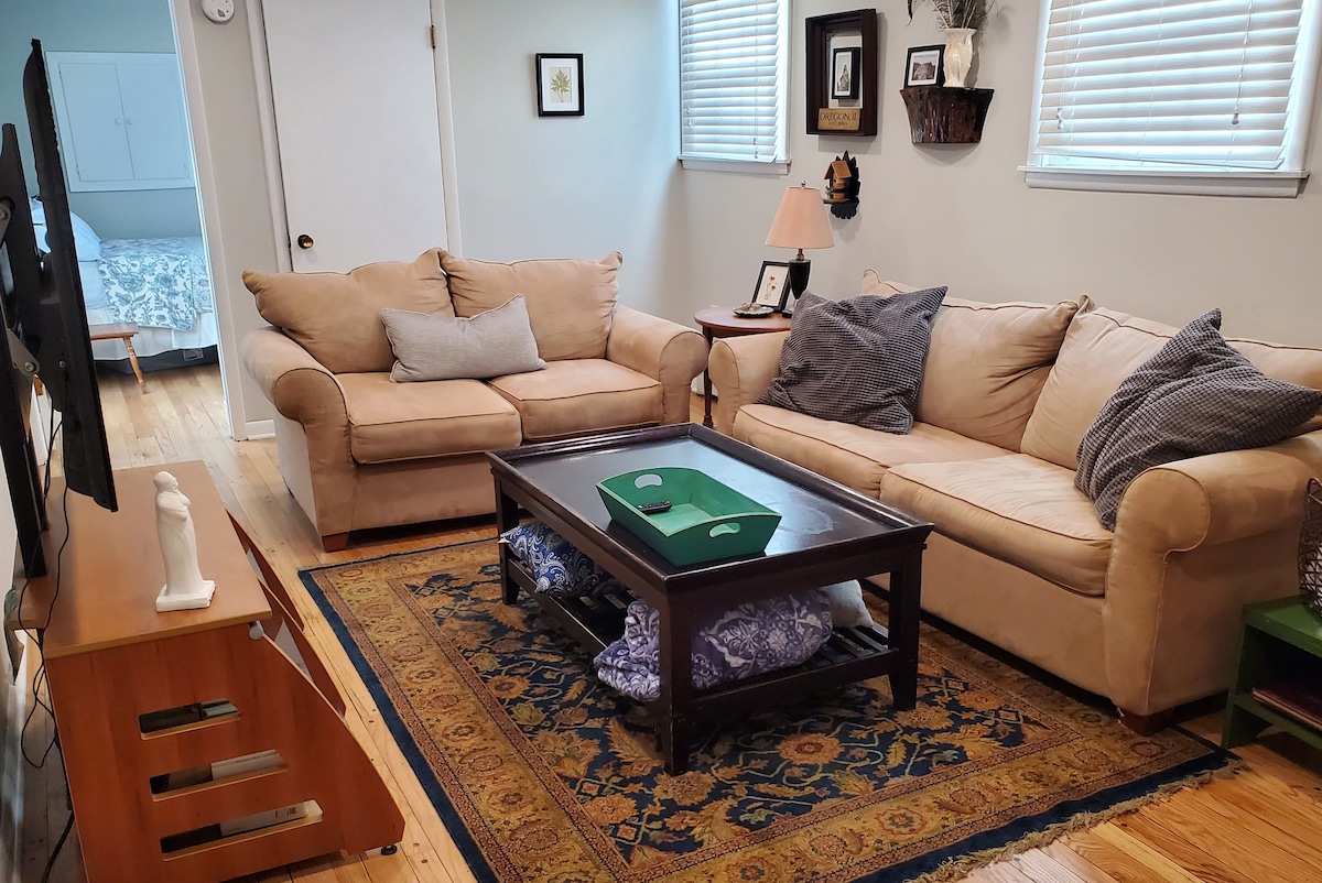 A comfortable living area features a beige sofa set arranged around a dark wooden coffee table. Soft pillows add to the seating comfort, while a decorative area rug brings warmth to the wood floors. Natural light filters through the window, brightening the space.