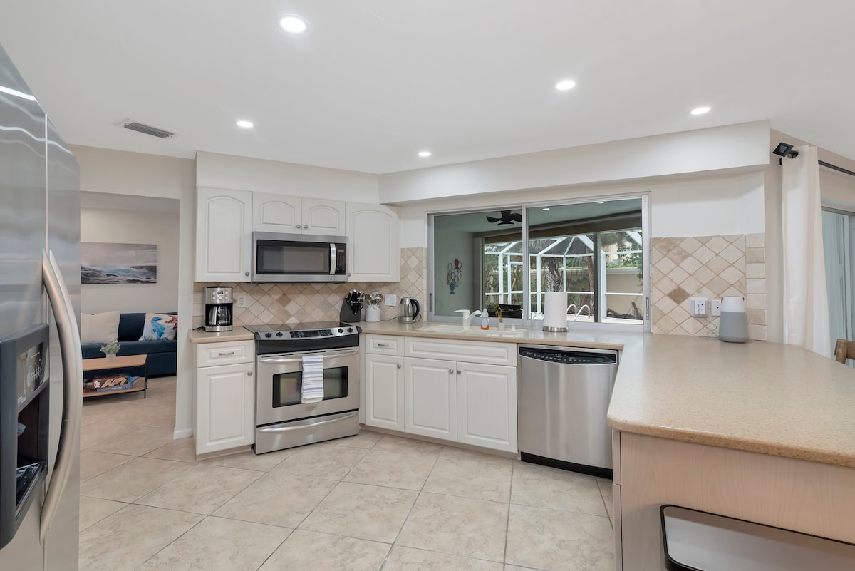 A modern kitchen is presented with stainless steel appliances, including a stove and refrigerator. Cream-colored cabinets complement the neutral tile flooring. Natural light floods in from a view of the enclosed patio, enhancing the spacious layout.