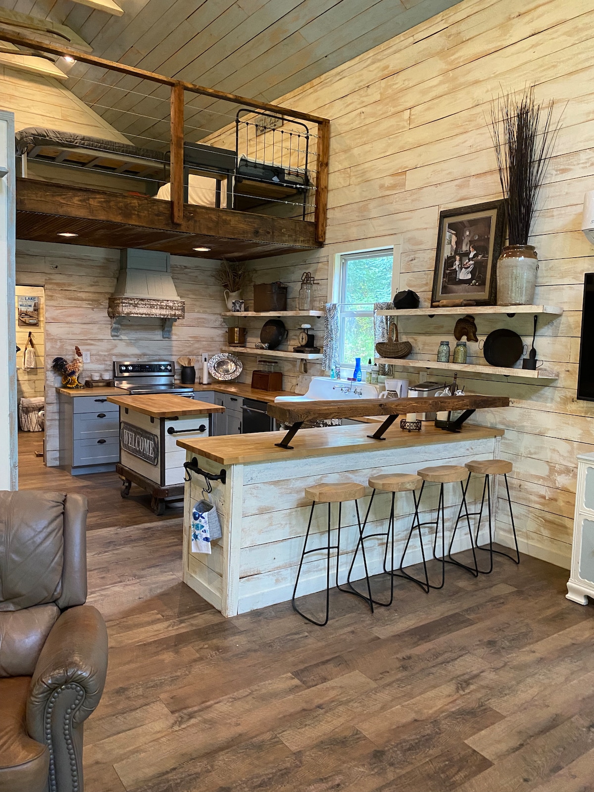 An open kitchen area features a wooden countertop with four metal bar stools. A rustic wooden shelving unit displays assorted dishware, while a stove and appliances are visible. An upstairs loft area is accessible by a staircase, showcasing additional sleeping arrangements.