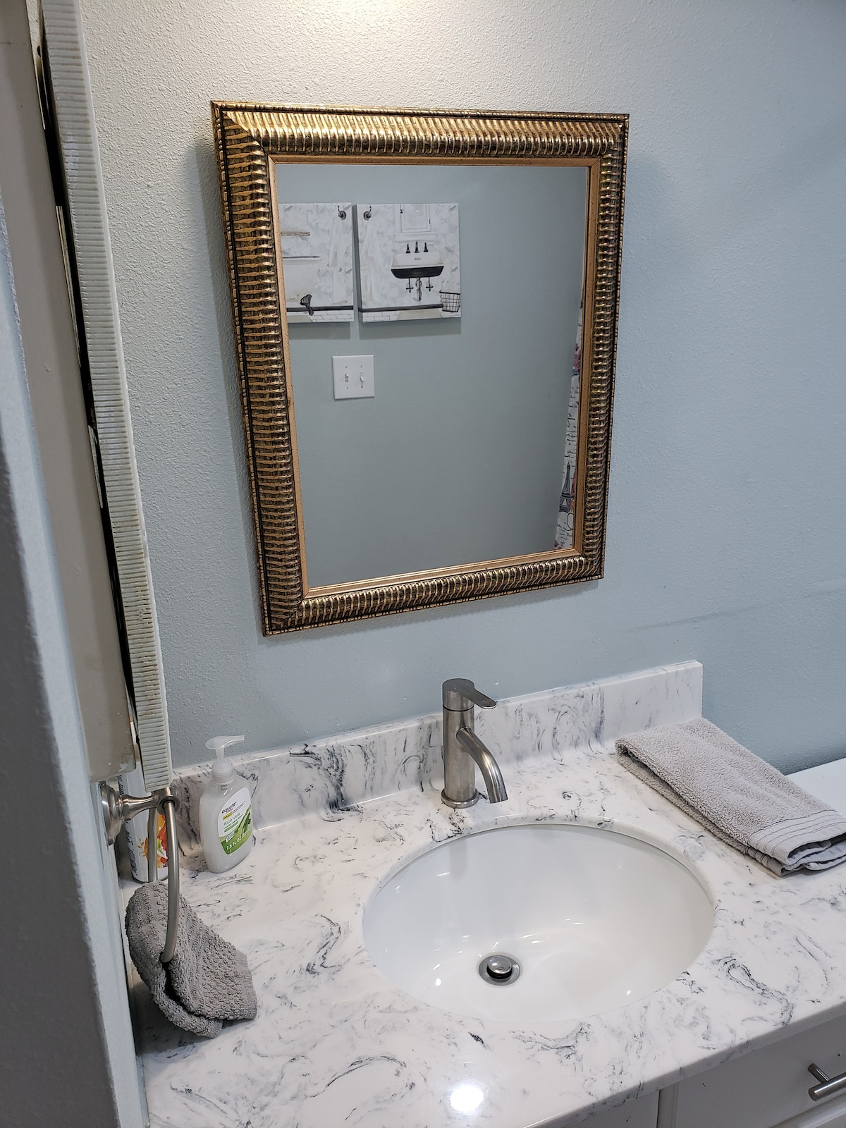 A modern bathroom sink area is presented with a marble countertop and a round white basin. A decorative framed mirror hangs above, reflecting the wall behind. Two neatly rolled towels are placed nearby, and a soap dispenser is positioned to the side.