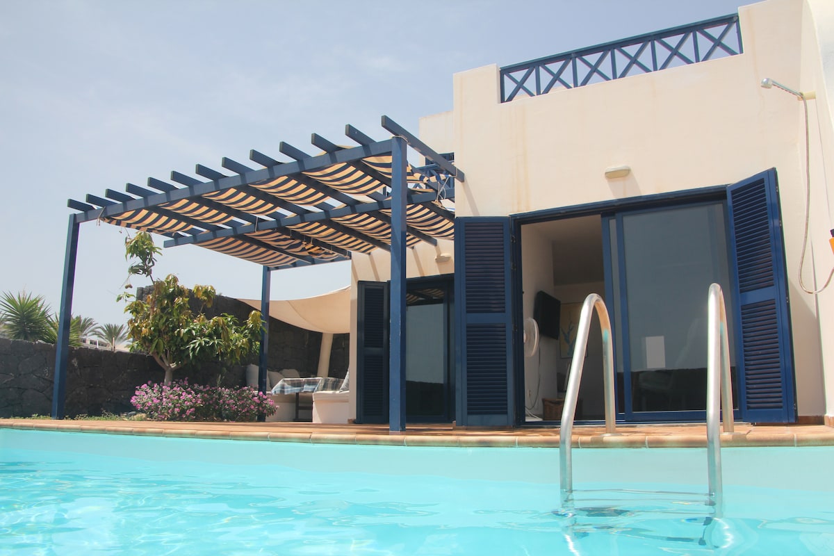 The image displays a villa with a designated shaded area supported by wooden beams, positioned next to a private pool. Large glass doors provide access to the interior, while landscaped greenery and colorful plants enhance the surrounding scenery.