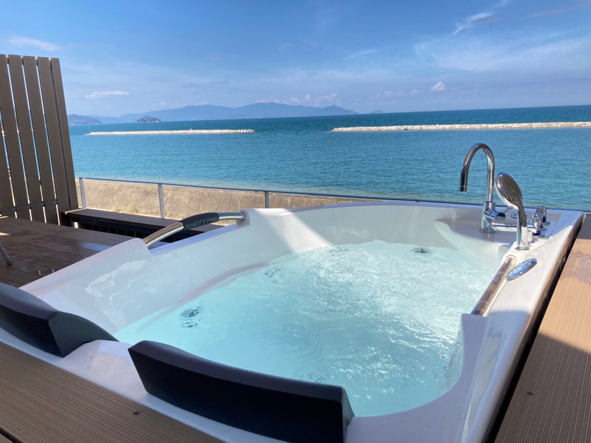 A private wood terrace features a spacious jacuzzi overlooking the ocean, where the gentle waves can be seen in the distance. The serene blue water with distant islands is visible, enhancing the tranquil environment.