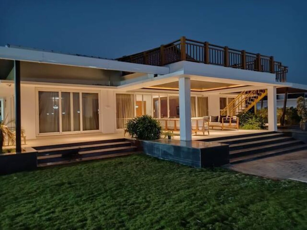The villa's exterior is illuminated at night, highlighting a spacious terrace above. Wide glass doors reveal an inviting indoor area, while steps lead to a well-maintained lawn, bordered by greenery.