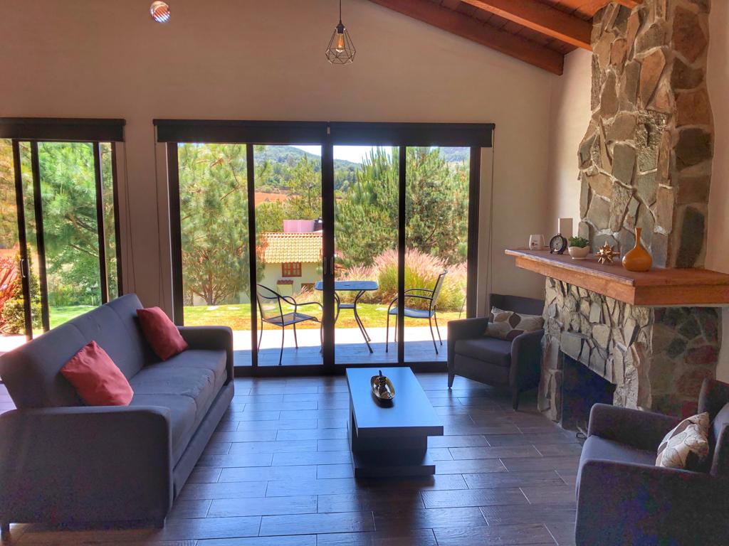 A living area is featured with large glass sliding doors, providing views of the surrounding greenery. A stone fireplace is positioned against a wooden wall, while two armchairs and a sofa with decorative cushions create a comfortable seating arrangement. A small coffee table is centered in the space.