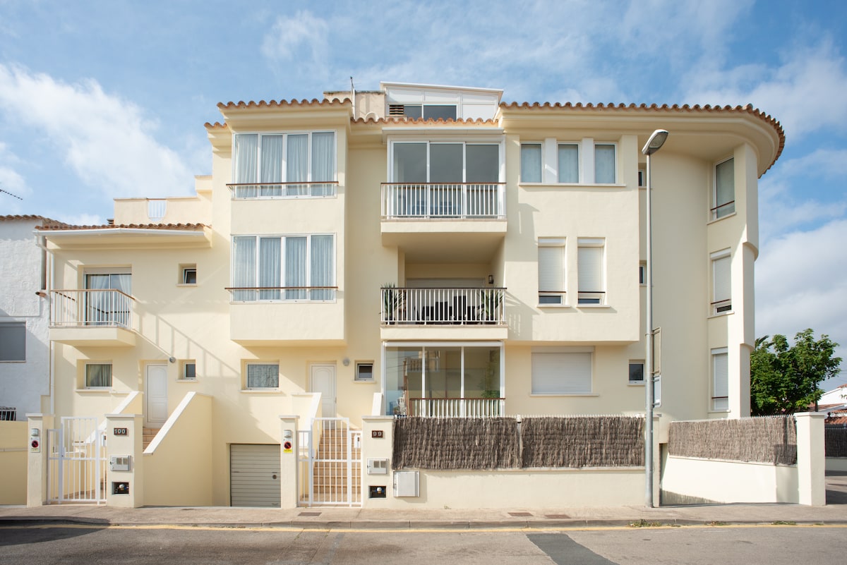 The exterior of a modern three-story building features a light-colored facade with multiple balconies. The design includes large windows and a gently sloping roof. A low fence surrounds the property, providing a sense of privacy while allowing views of the green landscaping.