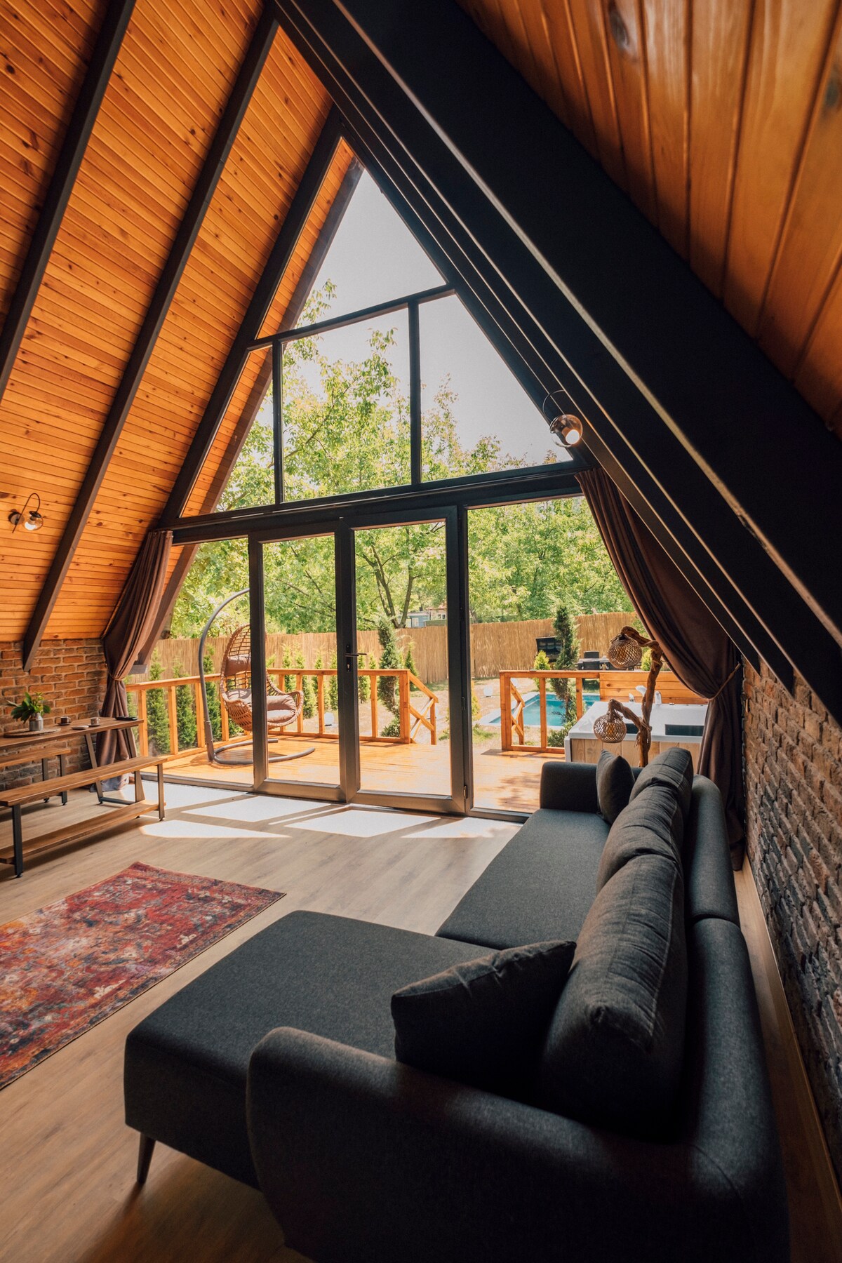 The image features a spacious living area with a large sectional sofa, situated beneath a high wooden ceiling. Expansive glass doors allow natural light to fill the room, offering views of the outdoor space, including a swimming pool and surrounding greenery.