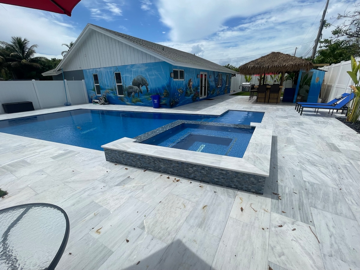 A heated swimming pool is surrounded by marble tiles, with a jacuzzi positioned nearby. A Tiki hut offers shade and seating. A mural depicting marine life adorns the home’s exterior, while lounge chairs invite relaxation under the clear sky.