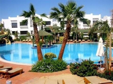 A large swimming pool is surrounded by palm trees and comfortable lounge chairs. A gazebo with a green roof is positioned near the water's edge, enhancing the tropical setting. Lush plants and flowers add a vibrant touch to the landscaped area.