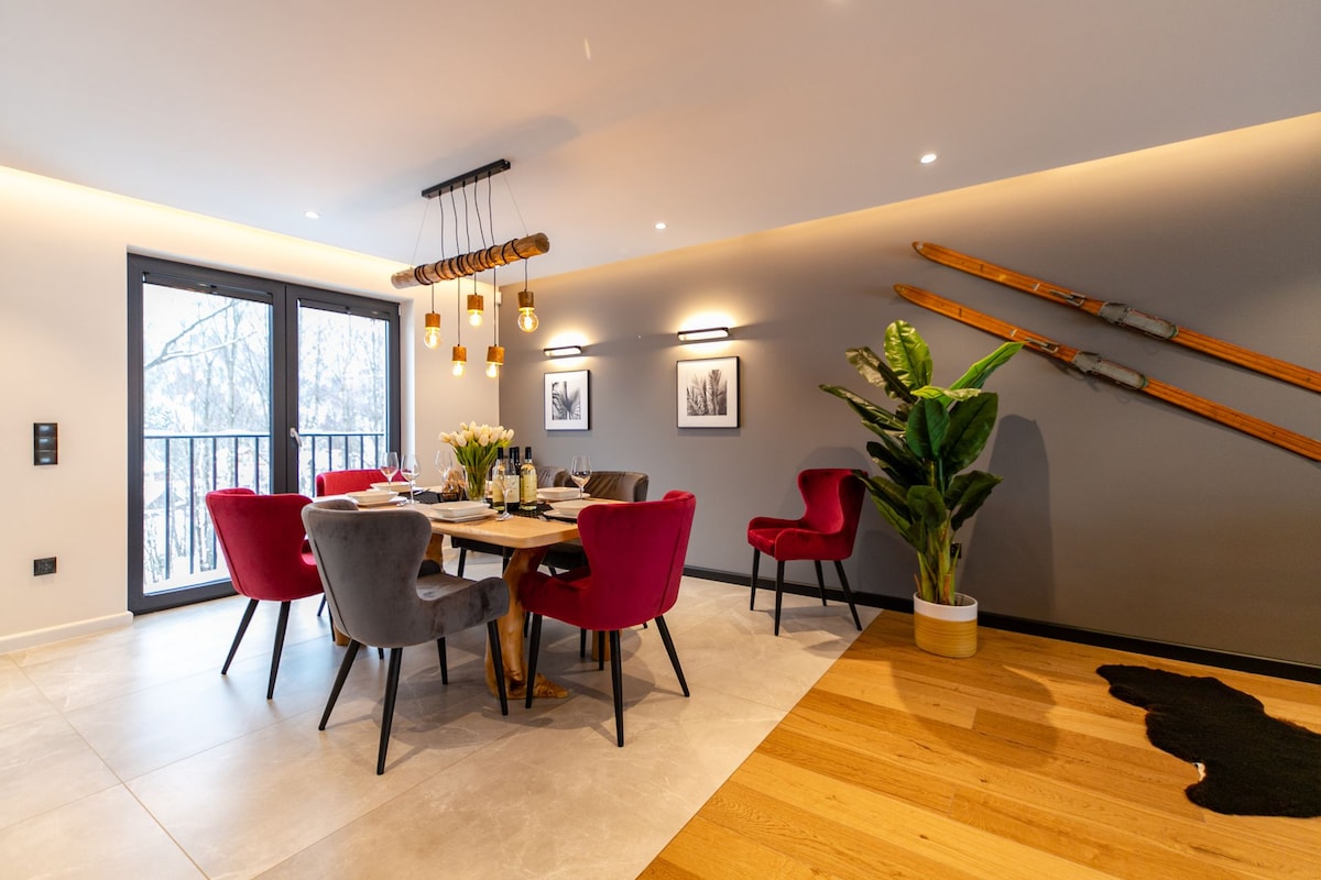 A stylish dining area is presented with a round table surrounded by four upholstered chairs in red and grey. Two large windows allow natural light to fill the space. Ski decorations are displayed on the wall, adding a cozy touch to the modern design.