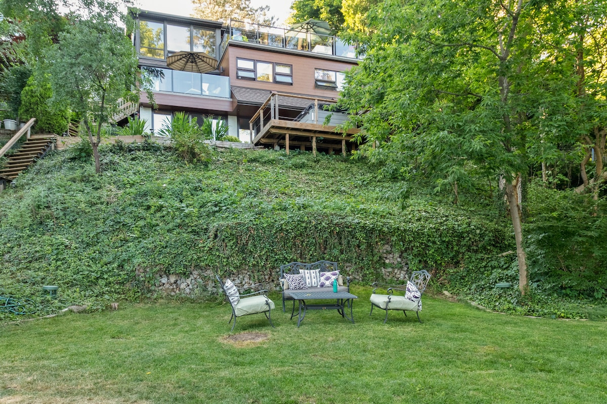 A grassy lawn features comfortable seating arrangements, including a table and chairs. The hillside villa is visible in the background, showcasing multiple outdoor decks among lush greenery. Surrounding trees provide a sense of privacy and tranquility in the outdoor space.