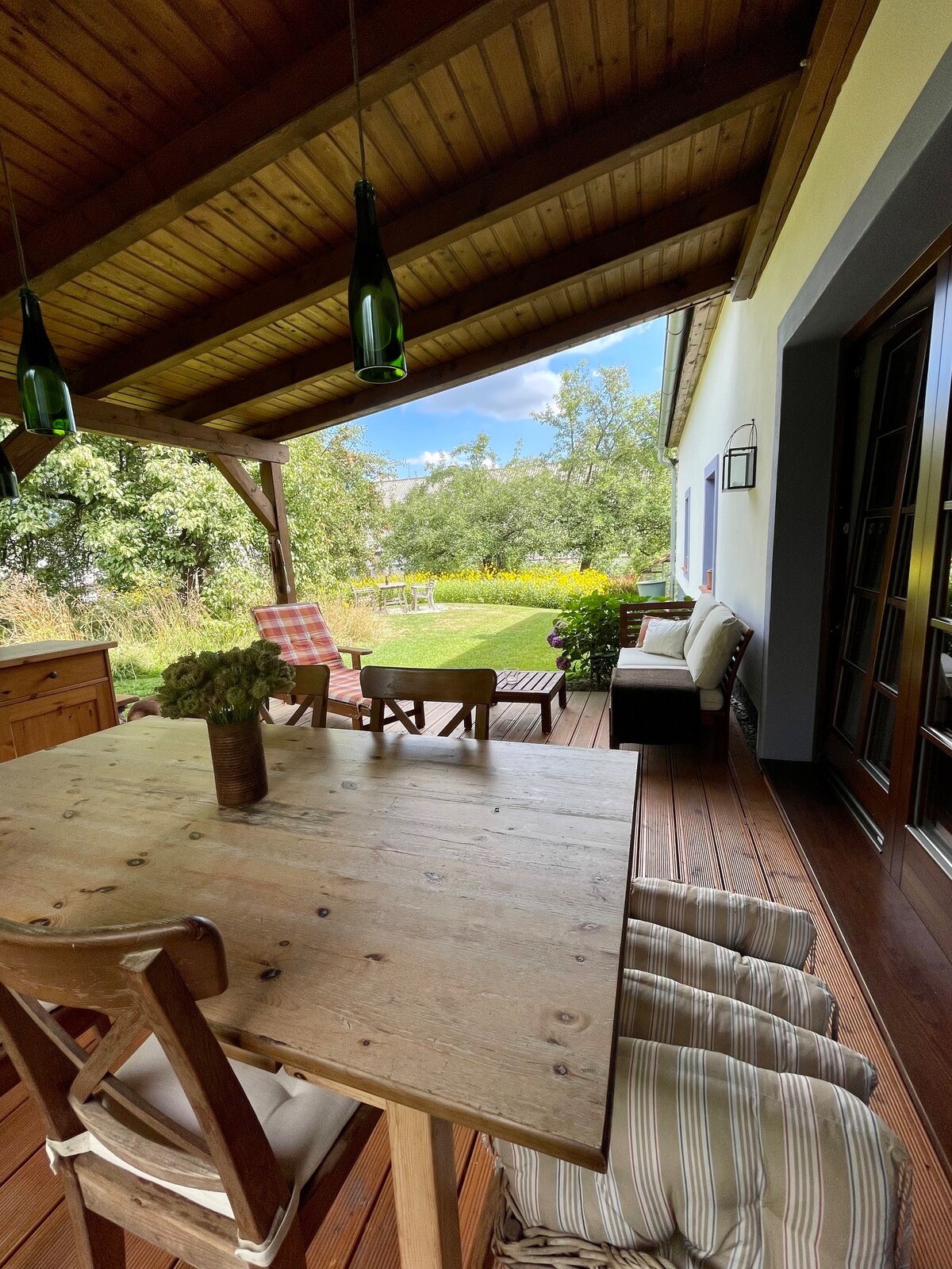 A covered outdoor patio features a wooden dining table surrounded by chairs with cushions. Lush greenery and colorful flowers are visible in the background, and comfortable seating is positioned nearby. Pendant lights hang from the wooden ceiling, adding a cozy touch.