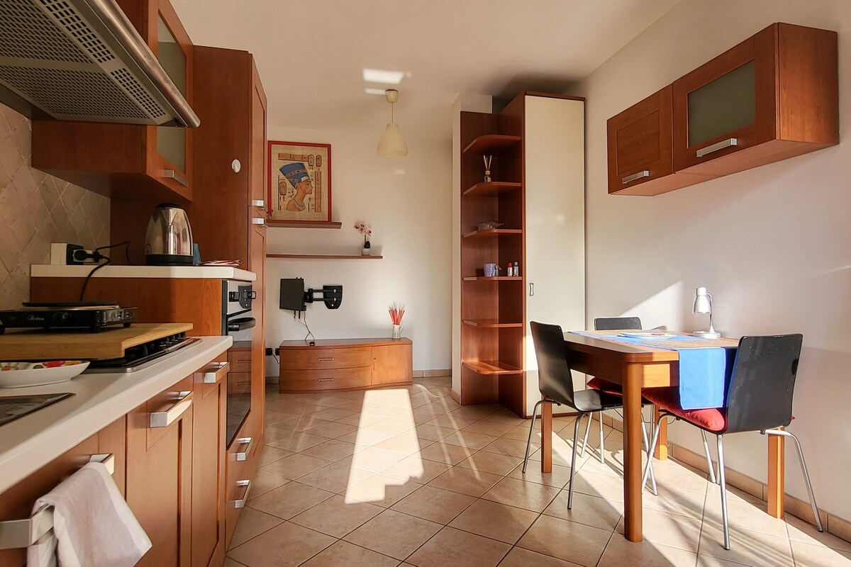 A modern kitchen space features warm wooden cabinetry and sleek appliances. A table with four chairs is positioned in the center, illuminated by natural light from a nearby window. Shelves display decorative items, and a framed artwork adds a touch of character to the wall.